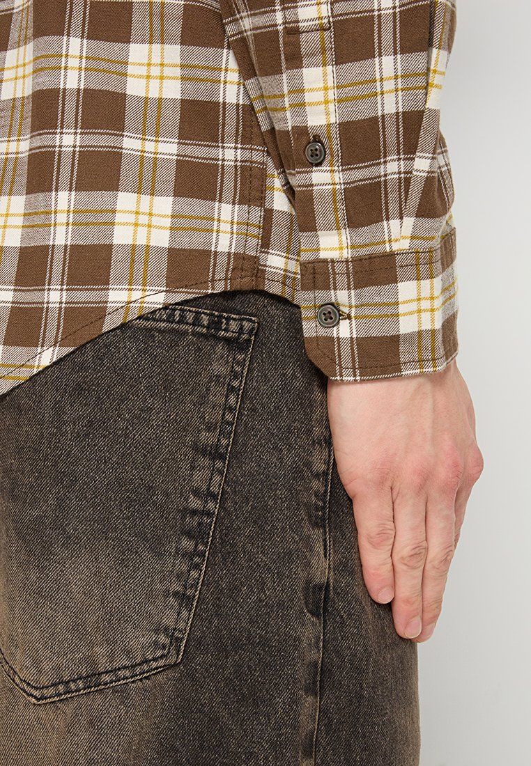 Braun kariertes Hemd mit gelben und weißen Akzenten, kombiniert mit abgenutzter schwarzer Denim, dabei sind die Knopfdetails an den Manschetten und die Handplatzierung zu sehen.