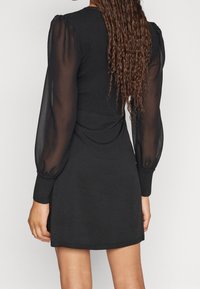 Schwarzes Kleid mit tailliertem, geripptem Oberteil und transparenten Puffärmeln. Glatte Textur mit elastischen Bündchen und einer lässigen, kurzen Länge.