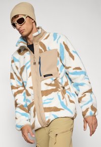 Giacca in pile bianca con motivo camouflage marrone e blu; presenta una tasca sul petto con zip color sabbia e un design a colletto aperto.