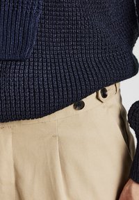 Maglione lavorato a maglia blu navale con texture a coste, abbinato a pantaloni beige con due bottoni scuri in vita e sottili righe di tessuto.