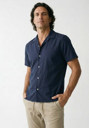 Hombre con cabello oscuro de longitud media, vestido con una camisa azul marino de manga corta con botones y pantalones beige, de pie con las manos en los bolsillos.