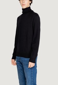 Maglione collo alto nero in tessuto testurizzato con polsini e orlo a coste, abbinato a jeans blu. Il maglione presenta un motivo sottile su tutta la superficie.