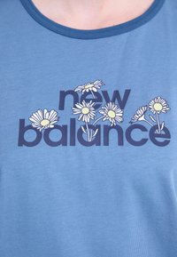 Mėlyna medvilninė marškinėliai su tamsiai mėlynu užrašu "new balance", aplink letteringą iliustruotos baltos ramunės.