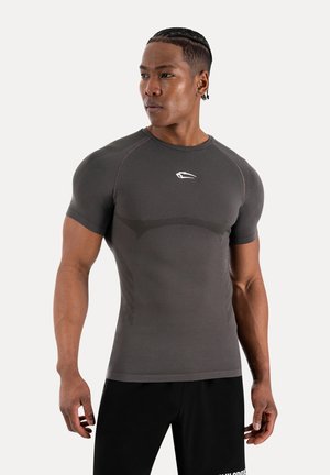 Athletischer Mann mit geflochtenen Haaren, der ein eng anliegendes dunkelgraues kurzärmeliges Shirt und schwarze Shorts trägt und nach links blickt, vor einem schlichten Hintergrund.