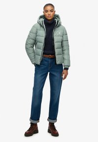Lysgrønne dunjakke med hætte, quiltet design og lynlåslukning. Sammen med en mørk sweater, blå jeans og brune snørestøvler.