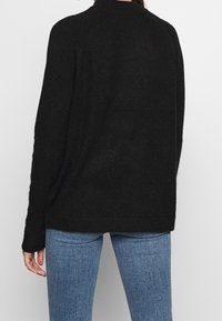 Pull noir en tricot avec un col montant, une coupe ample et un tissu texturé, associé à un jean bleu clair. Le matériau du pull semble doux.