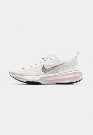 Chaussure de course Nike beige clair avec swoosh noir, semelle intermédiaire blanche, amorti rose et semelle extérieure noire, vue de côté sur fond blanc.