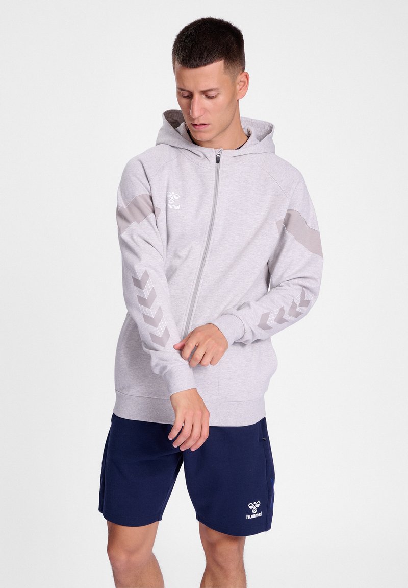Homme ajustant la manche d'un sweat à capuche zippé gris clair avec des bandes en chevrons, portant un short de sport bleu marine sur fond blanc.