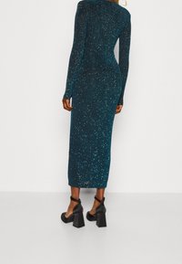 Türkises, langärmeliges, schimmerndes Strickkleid mit körperbetonter Silhouette, knielanger Länge und glatter Textur, kombiniert mit schwarzen Blockabsatzschuhen mit Knöchelriemen.