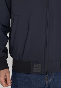Marineblaue Bomberjacke mit einem Frontreißverschluss, gerippten Bündchen und Saum sowie einem strukturierten Etikett an der Seite. Glattes, wasserabweisendes Material.
