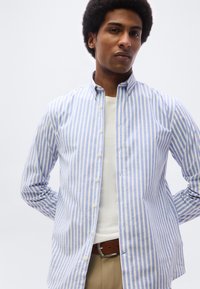 Άνδρας που φοράει ανοιχτό πουκάμισο με μπλε και λευκές ρίγες πάνω από λευκό μπλουζάκι, μπεζ παντελόνι και καφέ ζώνη, στέκεται με τα χέρια πίσω από την πλάτη.