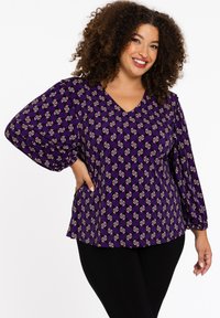 Lila Bluse mit V-Ausschnitt, puffigen Ärmeln und einem durchgehenden geometrischen Muster in Weiß und roten Akzenten. Kombiniert mit schwarzen Leggings.