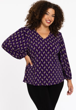 Blouse violette avec un décolleté en V, des manches bouffantes, et un motif géométrique total en blanc et rouge. Associée à des legging noirs.