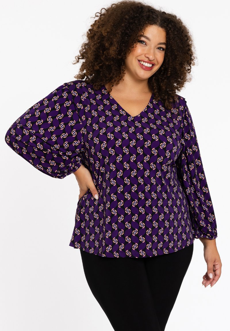 Lila Bluse mit V-Ausschnitt, puffigen Ärmeln und einem durchgehenden geometrischen Muster in Weiß und roten Akzenten. Kombiniert mit schwarzen Leggings.