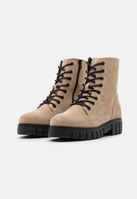 Botas de tobillo de ante beige con cordones negros. Presentan una punta redondeada y una cremallera lateral. Suela de goma negra gruesa con un patrón texturizado.