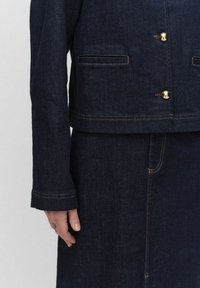 Veste en denim bleu marine avec un design court, ornée de boutons dorés, de poches latérales et de coutures orange contrastantes.