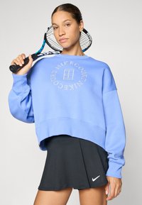 Lys blå cropped hettegenser med et sirkulært Nike-logo og svart plisserte skjørt med en liten hvit Nike swoosh.