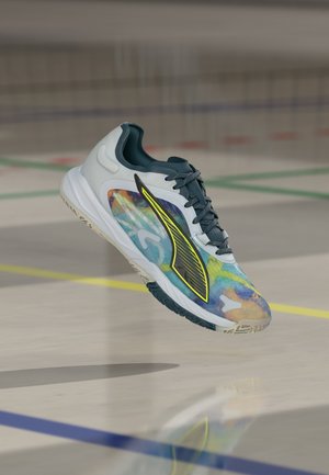 Lichtgrijze en meerkleurige sportschoen met zwart-geel logo, zwevend boven een gymzaalvloer met lijnen van een sportveld.