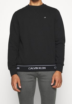 Mann trägt schwarzen Calvin Klein Pullover mit kleinem "ck" Logo auf der Brust und "Calvin Klein" Schriftzug am unteren Saum, kombiniert mit grauer Hose.