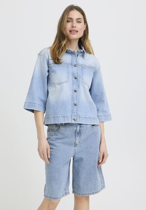 Femeie purtând o jachetă din denim bleu cu mâneci trei sferturi și pantaloni scurți asortați, din denim, până la genunchi, stând în fața unui fundal simplu.