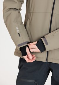 Beige vattentät jacka med en strukturerad yta, justerbara ärmslut med svarta och grå accenter, samt en dragkedja under kragen.