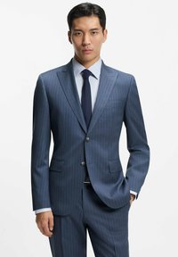 Granatowa marynarka w prążki z klapami szpiczastymi, dwoma guzikami i przednimi kieszeniami, w zestawie z jasną, prążkowaną koszulą i granatowym krawatem.