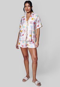 Mujer con camisa de manga corta abotonada y pantalones cortos a juego con estampados coloridos de temática marina, de pie contra un fondo liso.