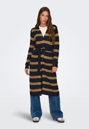 Femme debout portant un long cardigan en maille rayé noir et beige avec une ceinture noire, une chemise blanche, un jean bleu à jambes larges et des baskets blanches.