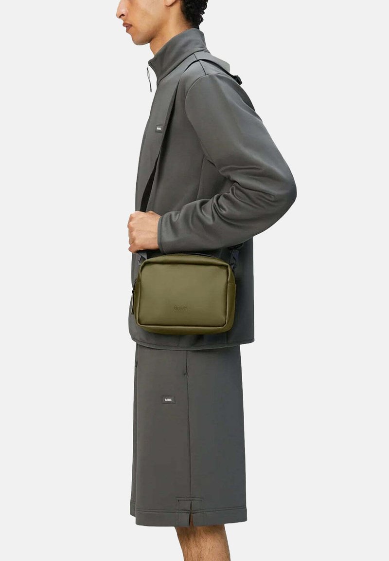 Borsa a tracolla verde oliva con una consistenza liscia, forma rettangolare e chiusura con zip. Indossata sotto un giubbotto grigio e pantaloni corti abbinati.