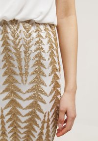 Jupe en sequins dorés présentant un motif triangulaire sur une base en tissu blanc. La texture est brillante, avec une taille élastique.