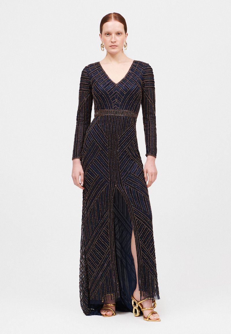 Lunga gown blu navy con profondo scollo a V, decorata con motivi geometrici oro e bronzo, e uno spacco laterale. Indossata con sandali dorati con cinturini.