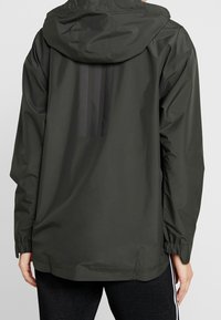 Mørkegrøn letvægtsjakke med hætte, der har en mat finish, elastiske manchetter og tre sorte lodrette striber på ryggen.