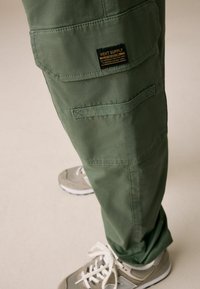 Pantalons cargo verts avec de multiples poches, arborant une étiquette en tissu portant la mention "NEXT SUPPLY". Portés avec des chaussures de sport grises, mettant en évidence les détails des lacets et de la semelle.