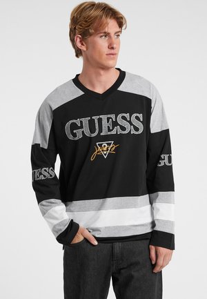 Maglietta a maniche lunghe nera, grigia e bianca con scollatura a V e logo "GUESS" insieme a delle strisce orizzontali. Presenta una vestibilità casual e un materiale in cotone morbido.