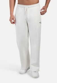 Homme portant un pantalon de survêtement blanc ample avec taille élastique et baskets blanches, se tenant debout devant un fond uni.