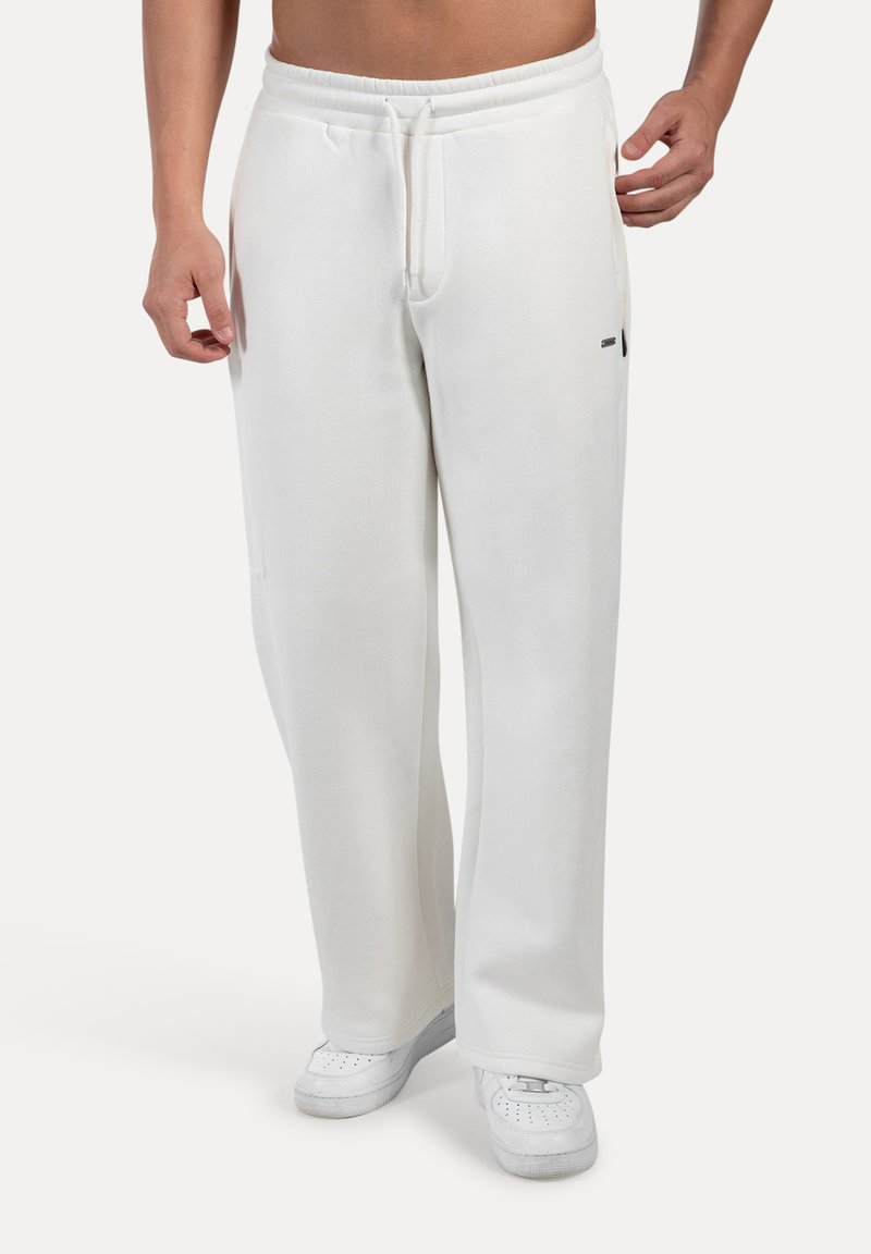 Homme portant un pantalon de survêtement blanc ample avec taille élastique et baskets blanches, se tenant debout devant un fond uni.