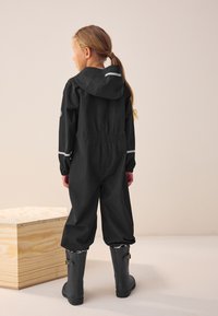 Black waterproof jumpsuit with a hood and reflective accents, elastic waist, and long sleeves, paired with gray rubber boots.