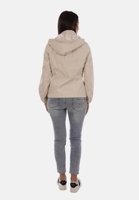 Donna in piedi, indossa una giacca beige con cappuccio, jeans aderenti grigi e sneakers bianche con dettagli neri, vista di schiena su sfondo semplice.