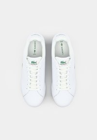 Sapatilhas de couro branco com bicos arredondados, atacadores planos e um destaque em logotipo verde. O interior apresenta palmilhas acolchoadas e uma textura suave.