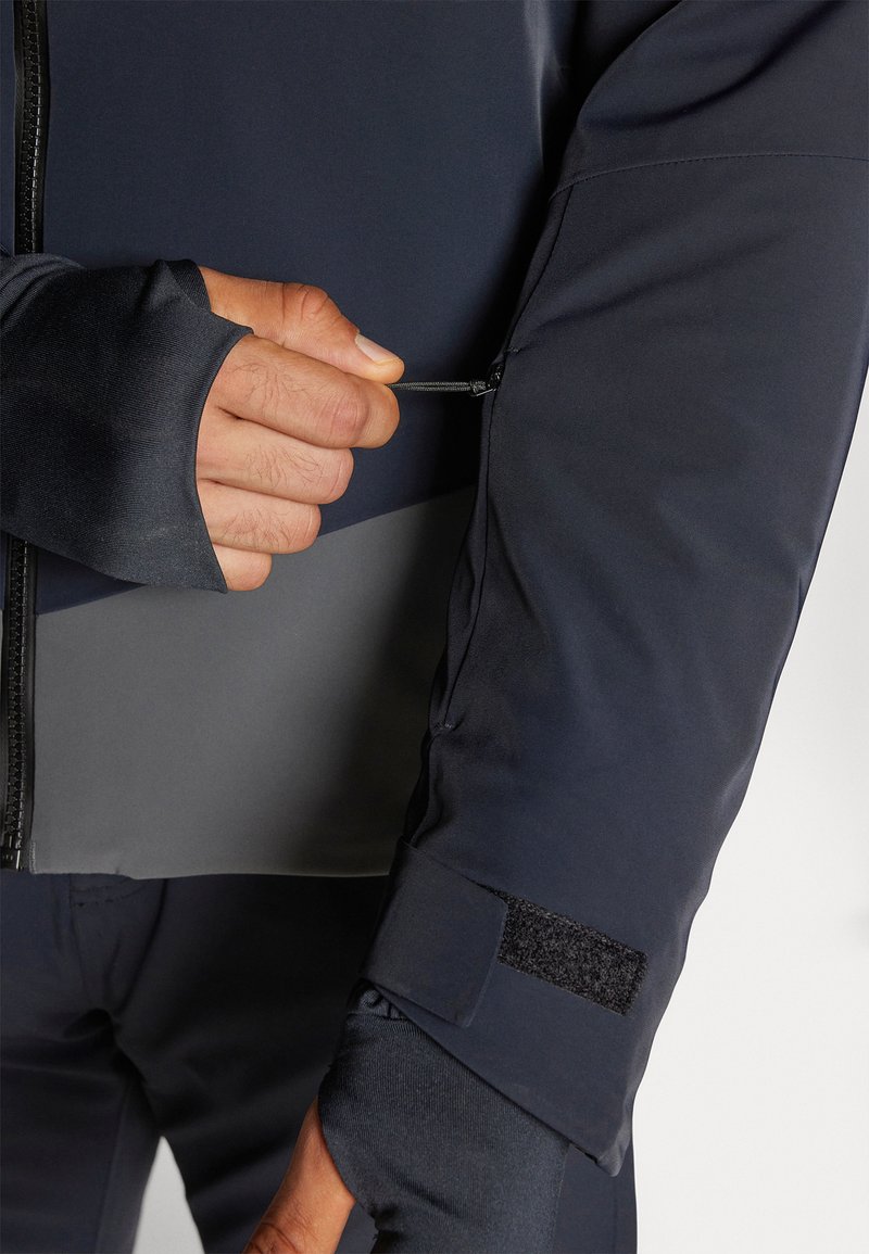 Mano che chiude con la cerniera una tasca della manica di una giacca blu scuro, con foro per il pollice e cinturino in velcro al polso, mostrando un primo piano dei dettagli dell'abbigliamento esterno.