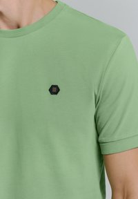 Lichtgroene T-shirt met korte mouwen van zacht materiaal, met een zwart hexagonaal logo op de linkerkant van de borst en een klassieke ronde halslijn.