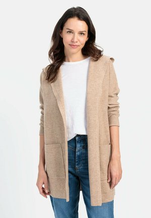 Beige Strickjacke mit Kapuze, offener Front, langen Ärmeln und zwei vorderen Taschen, getragen über einem weißen Hemd und blauen Jeans.
