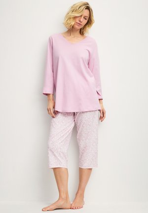Femme debout pieds nus portant un haut rose ample à manches longues et un pantalon court rose clair à motifs sur un fond uni.