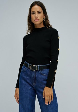 Jersey negro de punto con cuello alto, mangas ajustadas y detalles de botones dorados. Combinado con unos vaqueros de mezclilla azul de cintura alta y un cinturón negro.