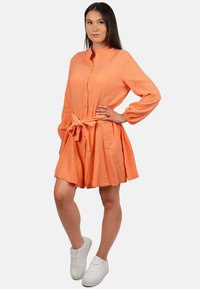 Abito arancione con bottoni, realizzato in tessuto testurizzato, dotato di maniche lunghe, vita regolabile con laccio e gonna svasata. Abbinato a sneakers bianche.