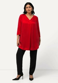 Rode tuniek met v-hals en driekwartmouwen, gecombineerd met zwarte rechte broek en zwarte puntige hakken, op een neutrale achtergrond.