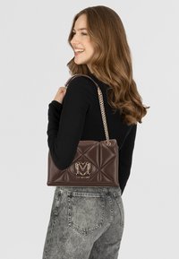 Borsa a tracolla in pelle marrone trapuntata con un accento del logo dorato, dotata di una tracolla a catena e una superficie testurizzata. Mostrata mentre viene tenuta sopra una maglia nera.