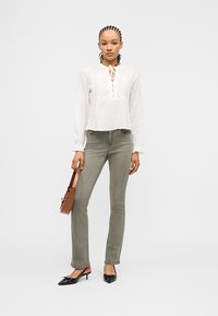 Γυναίκα με πλεγμένα μαλλιά, φοράει λευκό πουκάμισο με μακριά μανίκια, γκρι στενά τζιν, μαύρα χαμηλά τακούνια, κρατώντας ένα καφέ τσαντάκι στο πλάι της.