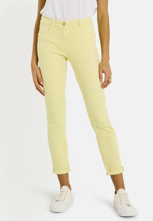 Mujer con vaqueros ajustados amarillo claro con bajos remangados y zapatillas blancas, de pie contra un fondo blanco liso.