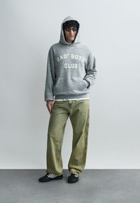 Hall hallipusa "BAD² BOYS CLUB" tekstiga, koos lahtiste soonikpükstega rohelises toonis ja mustade valgete triipudega kingadega. Aksessuaarideks päikeseprillid.